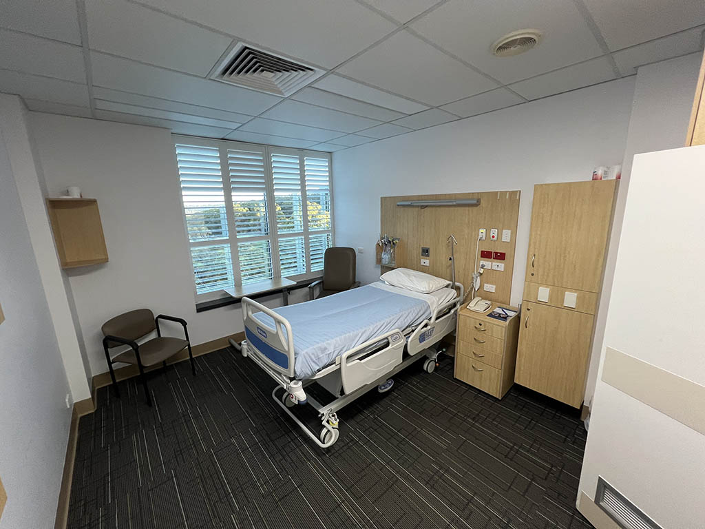 Hospital patient room with an adjustable bed, bedside table, and medical equipment along wooden cabinets by the window.