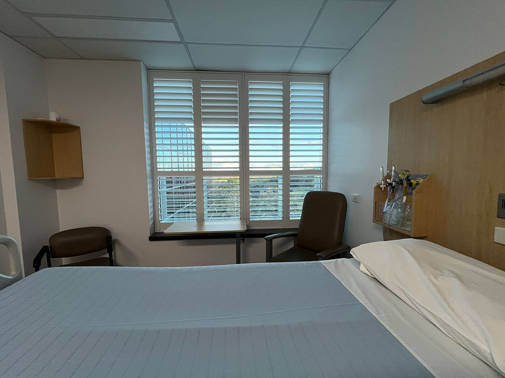 Hospital room with a patient bed made up in light blue sheets, a brown chair by a large window with blinds, and a small table at the window sill behind the chair.
